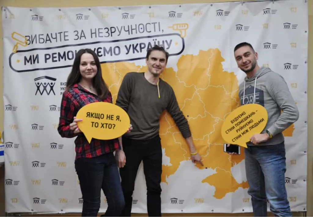 Троє волонтерів із мовними хмаринками «Якщо не я, то хто?» на тлі банера «Вибачте за незручності, ми ремонтуємо Україну» з мапою України.