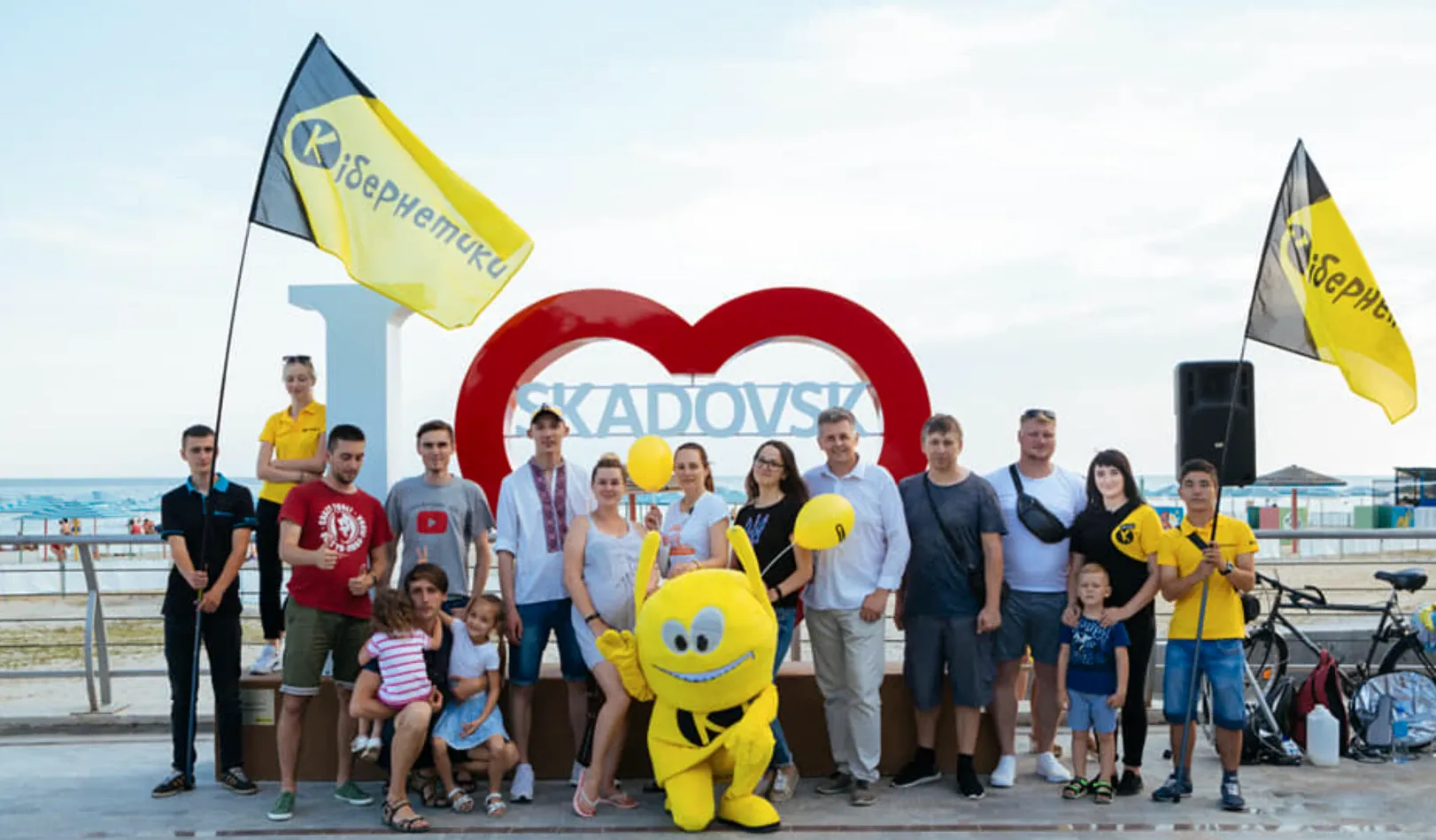 Велика група дорослих і дітей із маскотом біля інсталяції «I ♥ SKADOVSK» на набережній, із прапорами спільноти.