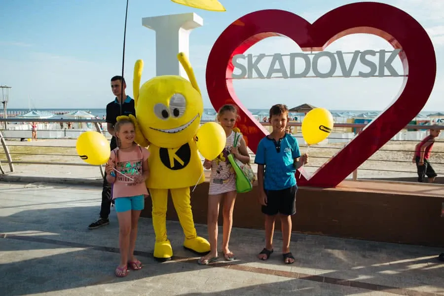 Троє дітей із жовтими кульками та маскот біля серця інсталяції «I ♥ SKADOVSK» на набережній.